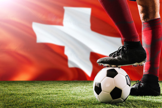 Close up legs of Switzerland football team player in red socks, shoes on soccer ball at the free kick or penalty spot playing on grass.
