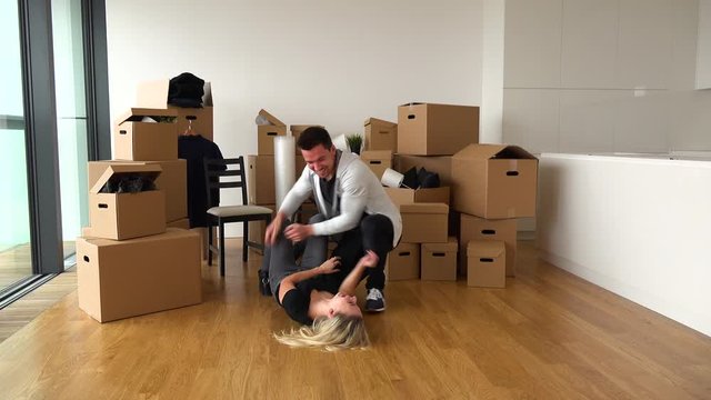 A playful moving couple in a new apartment, the woman lies on the floor, the man tickles her - piles of cardboard boxes in the background