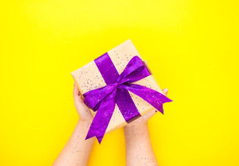 Child's hands holding giftbox tied with purple color ribbon on bright yellow background with glitter. Flat lay style.