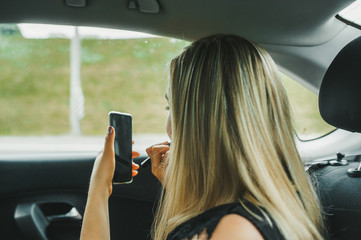 Blonde woman sitting in car with cellphone and cosmetics. Back view.