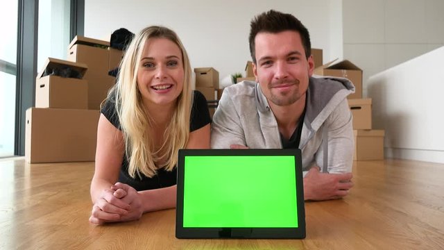 A smiling moving couple lies on the floor of an empty apartment and talks to the camera - tablet with green screen in the foreground - cardboard boxes in the background