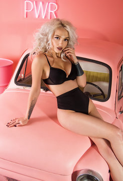 young glamour girl with platinum blonde long hair with perfect body in black minimal underwear is sitting on pink retro car and looking flirty on pink wall background, pin-up concept, free space