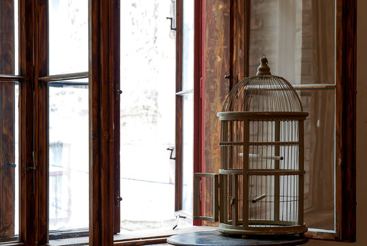 silhouette of an empty vintage bird cage standing in a window sill with wooden window frames