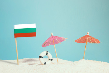 Miniature flag of Bulgaria on beach with colorful umbrellas and life preserver. Travel concept, summer theme.