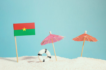 Miniature flag of Burkina Faso on beach with colorful umbrellas and life preserver. Travel concept, summer theme.