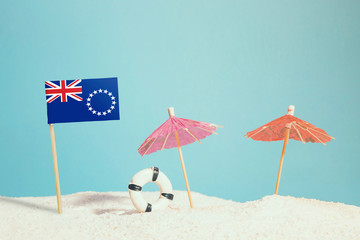 Miniature flag of Cook Islands on beach with colorful umbrellas and life preserver. Travel concept, summer theme.