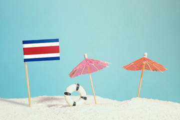 Miniature flag of Costa Rica on beach with colorful umbrellas and life preserver. Travel concept, summer theme.