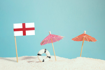 Miniature flag of England on beach with colorful umbrellas and life preserver. Travel concept, summer theme.