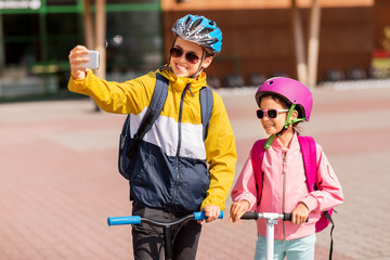education, childhood and people concept - happy school children in helmets with backpacks riding scooters and taking selfie by smartphone outdoors