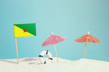 Miniature flag of French Guiana on beach with colorful umbrellas and life preserver. Travel concept, summer theme.
