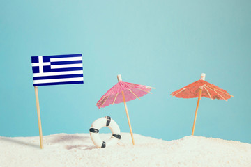 Miniature flag of Greece on beach with colorful umbrellas and life preserver. Travel concept, summer theme.