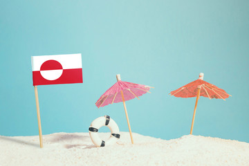 Miniature flag of Greenland on beach with colorful umbrellas and life preserver. Travel concept, summer theme.