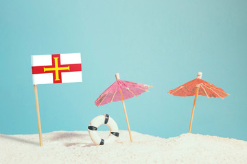 Miniature flag of Guernsey on beach with colorful umbrellas and life preserver. Travel concept, summer theme.