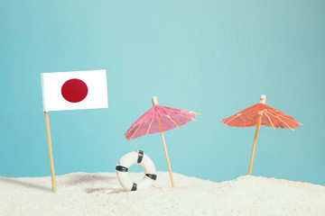 Miniature flag of Japan on beach with colorful umbrellas and life preserver. Travel concept, summer theme.