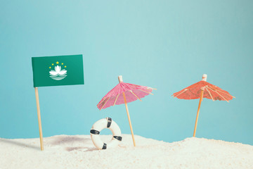 Miniature flag of Macao on beach with colorful umbrellas and life preserver. Travel concept, summer theme.
