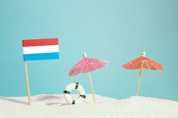 Fototapeta na wymiar Miniature flag of Luxembourg on beach with colorful umbrellas and life preserver. Travel concept, summer theme.