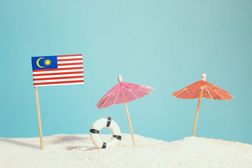 Miniature flag of Malaysia on beach with colorful umbrellas and life preserver. Travel concept, summer theme.