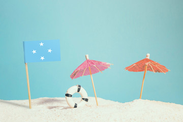 Miniature flag of Micronesia on beach with colorful umbrellas and life preserver. Travel concept, summer theme.