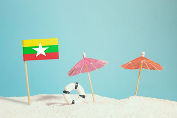 Miniature flag of Myanmar on beach with colorful umbrellas and life preserver. Travel concept, summer theme.