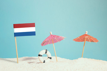 Miniature flag of Netherlands on beach with colorful umbrellas and life preserver. Travel concept, summer theme.