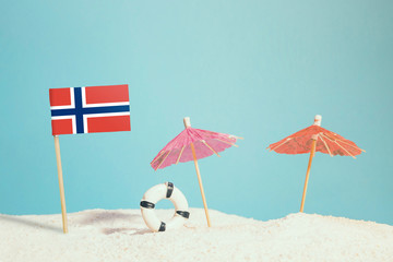 Miniature flag of Norway on beach with colorful umbrellas and life preserver. Travel concept, summer theme.