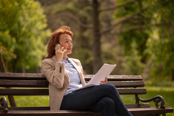 one mature woman, speaking over phone, while looking at papers in park.