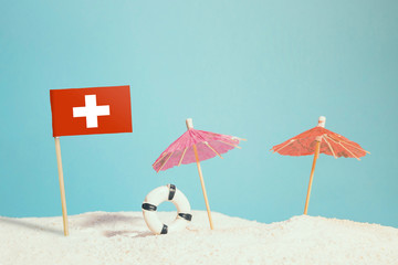 Miniature flag of Switzerland on beach with colorful umbrellas and life preserver. Travel concept, summer theme.