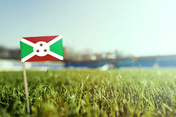 Miniature stick Burundi flag on green grass, close up sunny field. Stadium background, copy space for text.