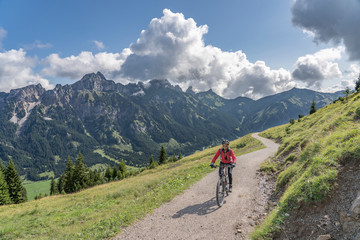 Fototapeta na wymiar nice and active senior woman, riding her e-mountain bike in the Tannheim valley , Tirol, Austria, with the village of Tannheim and famous summits Gimpel and Rote Flueh