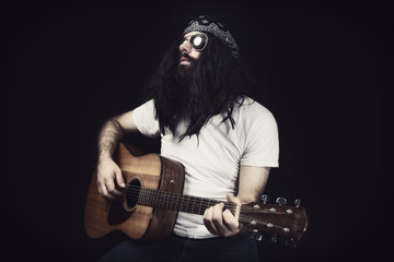Hippie with long hair, a bandanna and white shirt playing an acoustic guitar. Set against a black...