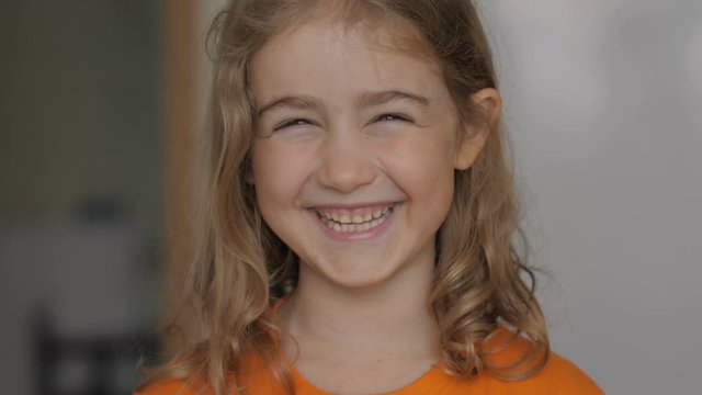 Little girl looks out from behind balloon and laughs happily. Close up portrait of a happy little child bursting in laugh. Child girl blonde with blue eyes and laughs indoors. Closeup. Slow Motion.
