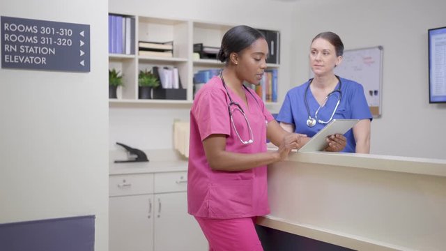 Two nurses w/ stethoscopes at nurses station review chart, camera move left