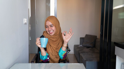 portrait of Happy muslim woman with hijab sitting in kitchen holding cup of tea of coffee enjoying her relaxing free time