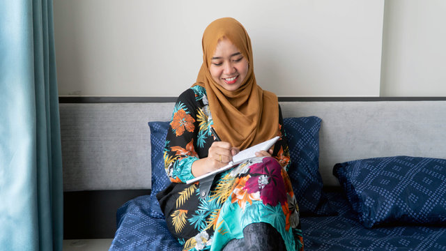 Cropped shot of Hijab woman sits crossed legs, wears pyjamas, makes notes in diary