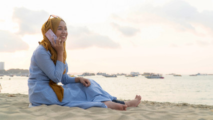 portrait of young muslim asia woman with hijab and blue dress using mobile phone and do a phone call on a beach