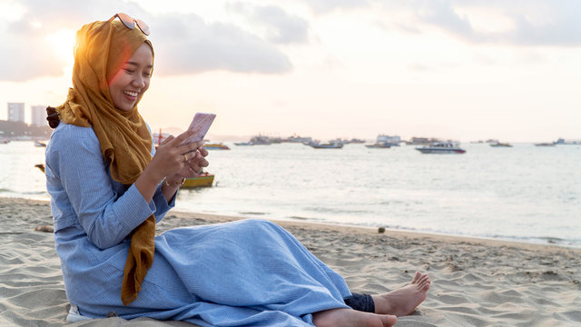 portrait of young muslim asia woman with hijab and blue dress using mobile phone on a beach on sunset in summer