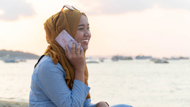 portrait of young muslim asia woman with hijab and blue dress using mobile phone and do a phone call on a beach