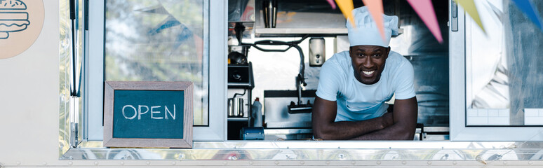 panoramic shot of happy african american man in chef uniform smiling from food truck