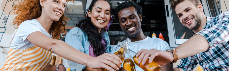 panoramic shot of happy multicultural friends clinking bottles with beer near food truck