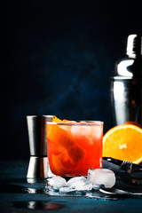 Garibaldi alcoholic cocktail with red bitter, orange juice, zest and ice. Dark blue background, bar tools, selective focus