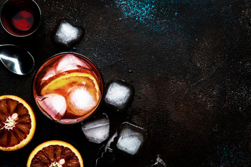 Negroni alcoholic cocktail beverage with dry gin, red vermouth and red bitter, bloody orange slice and ice cubes. Brown bar counter background, steel bar tools, top view, with copy space