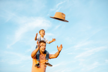 Portrait of a happy father with young son riding on the shoulders, playign with hat on the blue sky background outdoors. Concept of a happy family on a summer activity