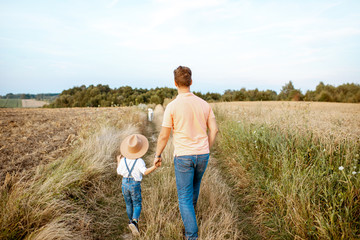 Father and young son walking keeping hands together on the field during the summer activity, back view