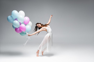 young, beautiful ballerina dancing with festive balloons on grey background