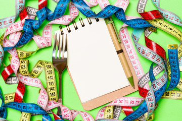 Group of colorful measure tapes, pen, open notebook and fork on green background with empty space for your idea. Top view of healthy lifestyle concept