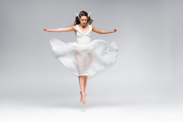 young attractive ballerina in white dress jumping while dancing on grey background