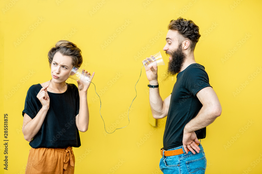 Wall mural Man and woman talking with string phone made of cups on the yellow background. Concept of communication