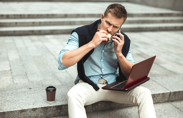 Obraz premium young handsome office worker having conversation while biting his sandwich and drinking coffee for lunch during break sitting on the stairs outside with laptop
