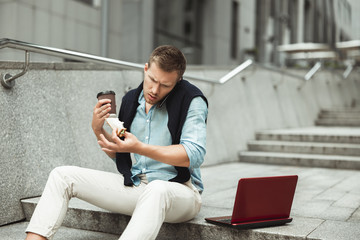 young handsome office worker having business conversation while eating his sandwich and drinking coffee for lunch during break sitting on the stairs outside with laptop