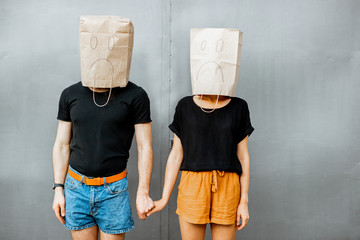 Portrait of a young couple with paper bags on their heads, keeping hands together on the grey wall...